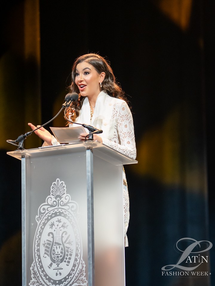 A speaker in a white outfit addresses the audience from the awards podium